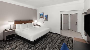 A hotel room with a bed, bedside tables, a desk, a chair, and a TV. There are two doors and a small window.