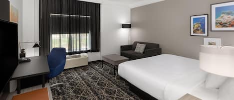Habitación de hotel con cama, escritorio, silla, sofá, lámpara y ventana con cortinas.
