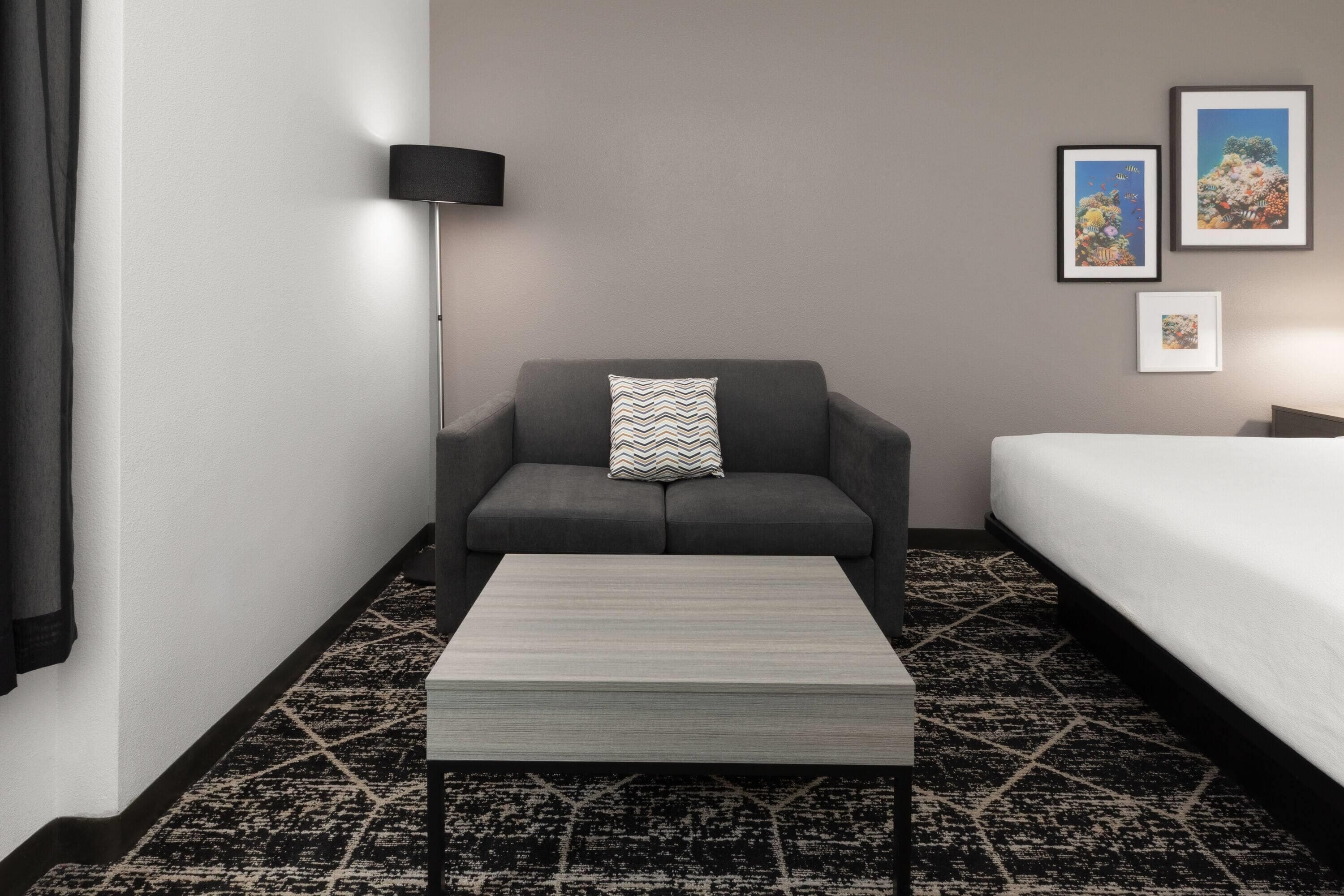 A hotel room with a grey sofa, a small table, a bed with white bedding, a floor lamp, and two framed pictures on the wall.