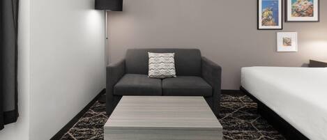 A hotel room with a grey sofa, a small table, a bed with white bedding, a floor lamp, and two framed pictures on the wall.
