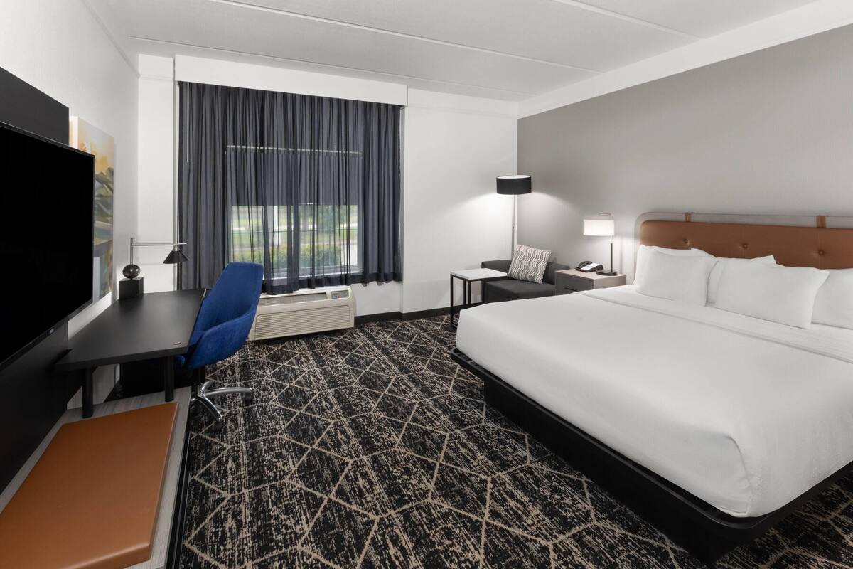 Een hotelkamer met een groot bed, een bureau met stoel, een televisie, een lamp en een raam met gordijnen.