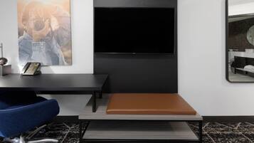 A modern hotel room with a large flat-screen TV mounted on the wall, a desk with a chair, a bench, and a patterned carpet.