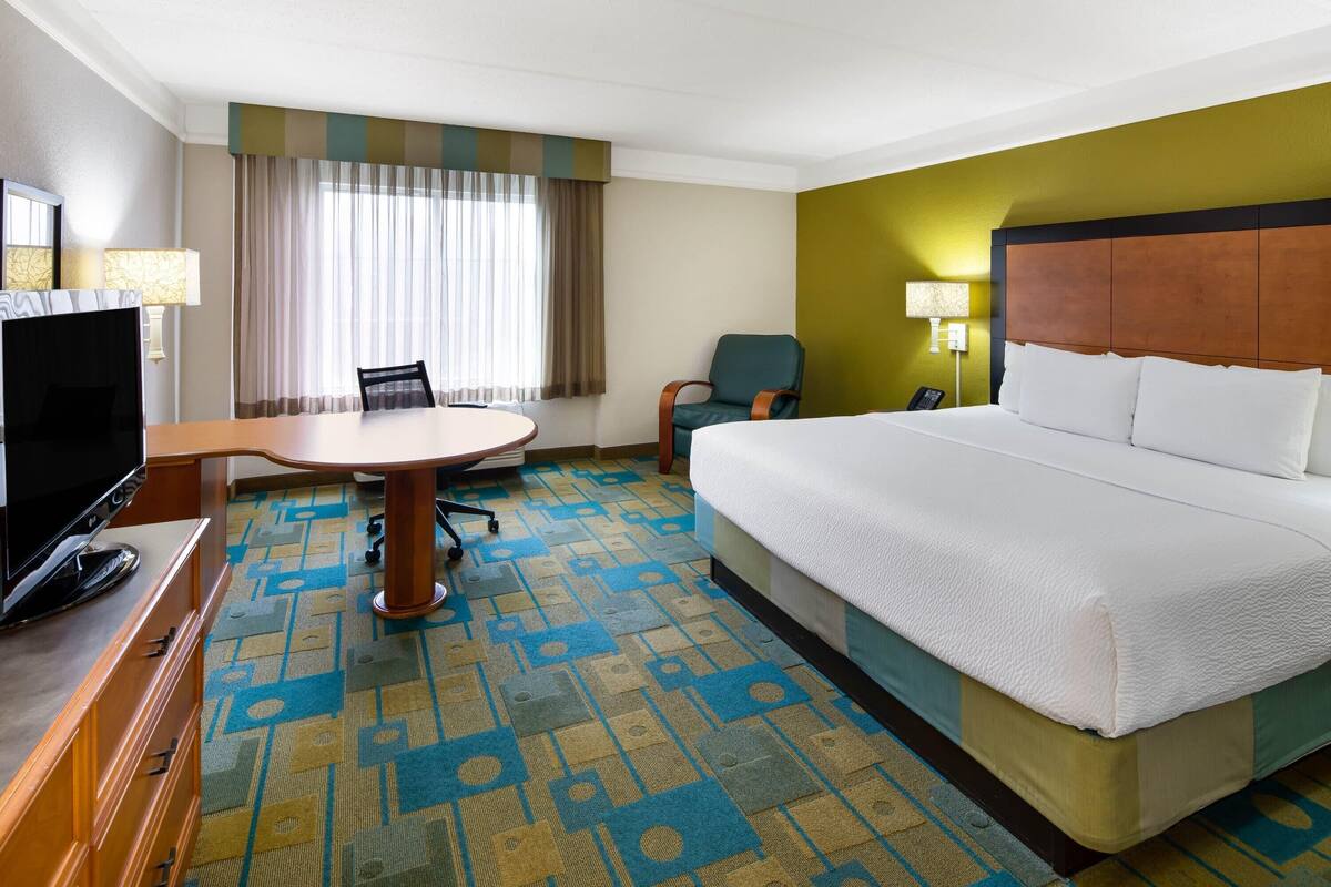 Ein Hotelzimmer mit einem großen Bett, einem Schreibtisch mit Fernseher, einem Sessel und einem Fenster mit Vorhängen.