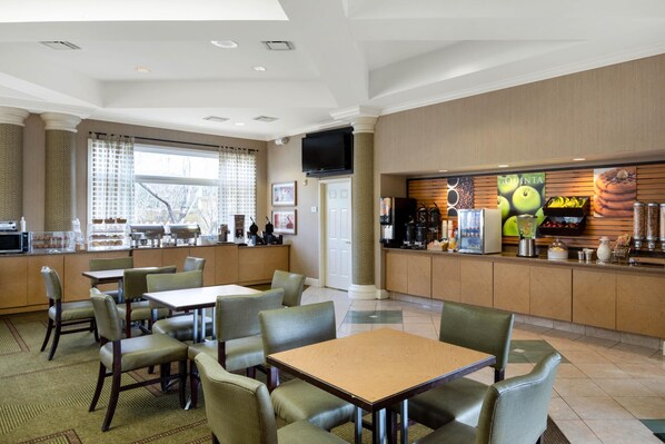 Un comedor de hotel con varias mesas y sillas, una barra de cocina con estación de café, y un amplio ventanal con persianas.