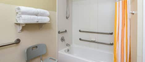 A bathroom with a white bathtub, a shower with a glass door, a grab bar, a towel rack, and a wheelchair-accessible bench.