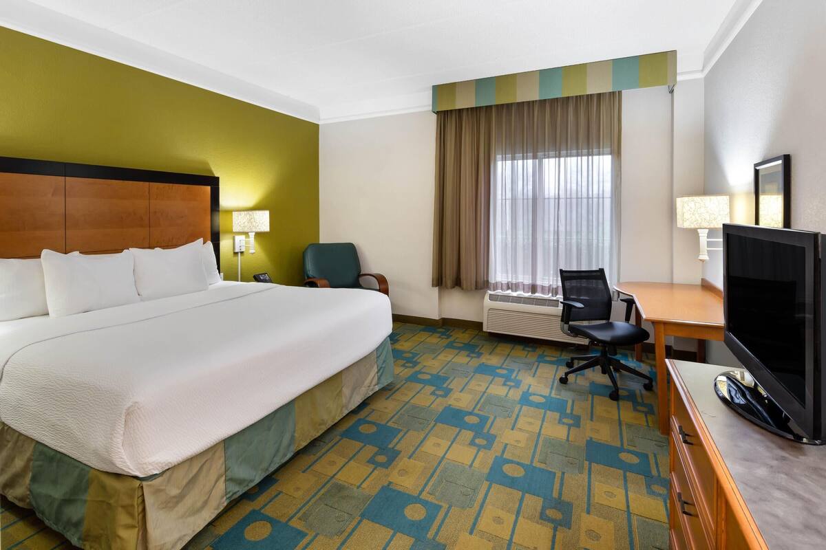 Ein Hotelzimmer mit einem großen Bett, einem Schreibtisch mit Fernseher, einem Sessel und einem Fenster mit Vorhängen.