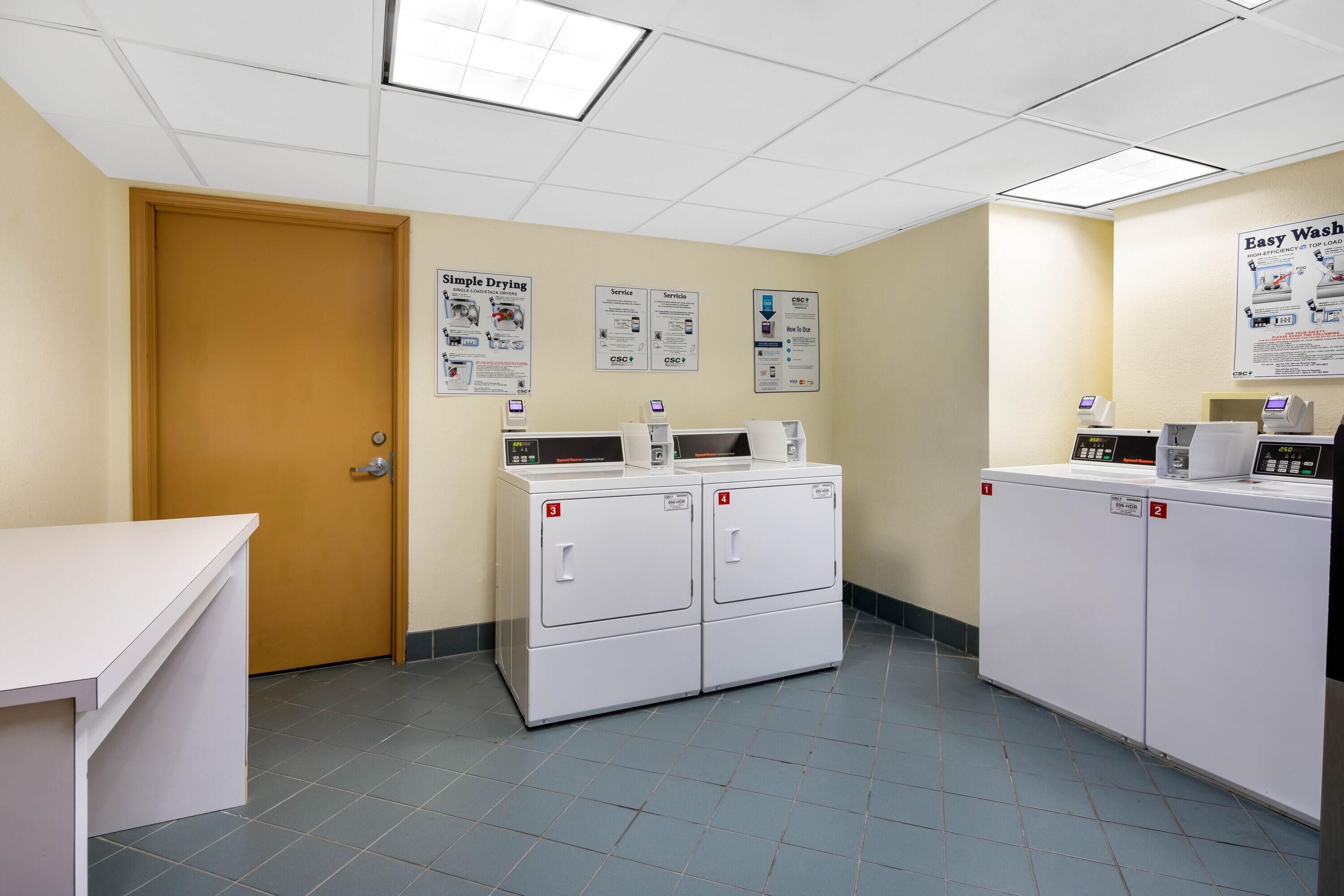 A laundry room with four washing machines, a drying machine, and a white counter. There are posters on the wall and a door to the left.