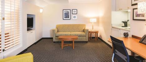 A hotel room with a yellow sofa, a wooden coffee table, a TV, a lamp, and a desk with a chair.