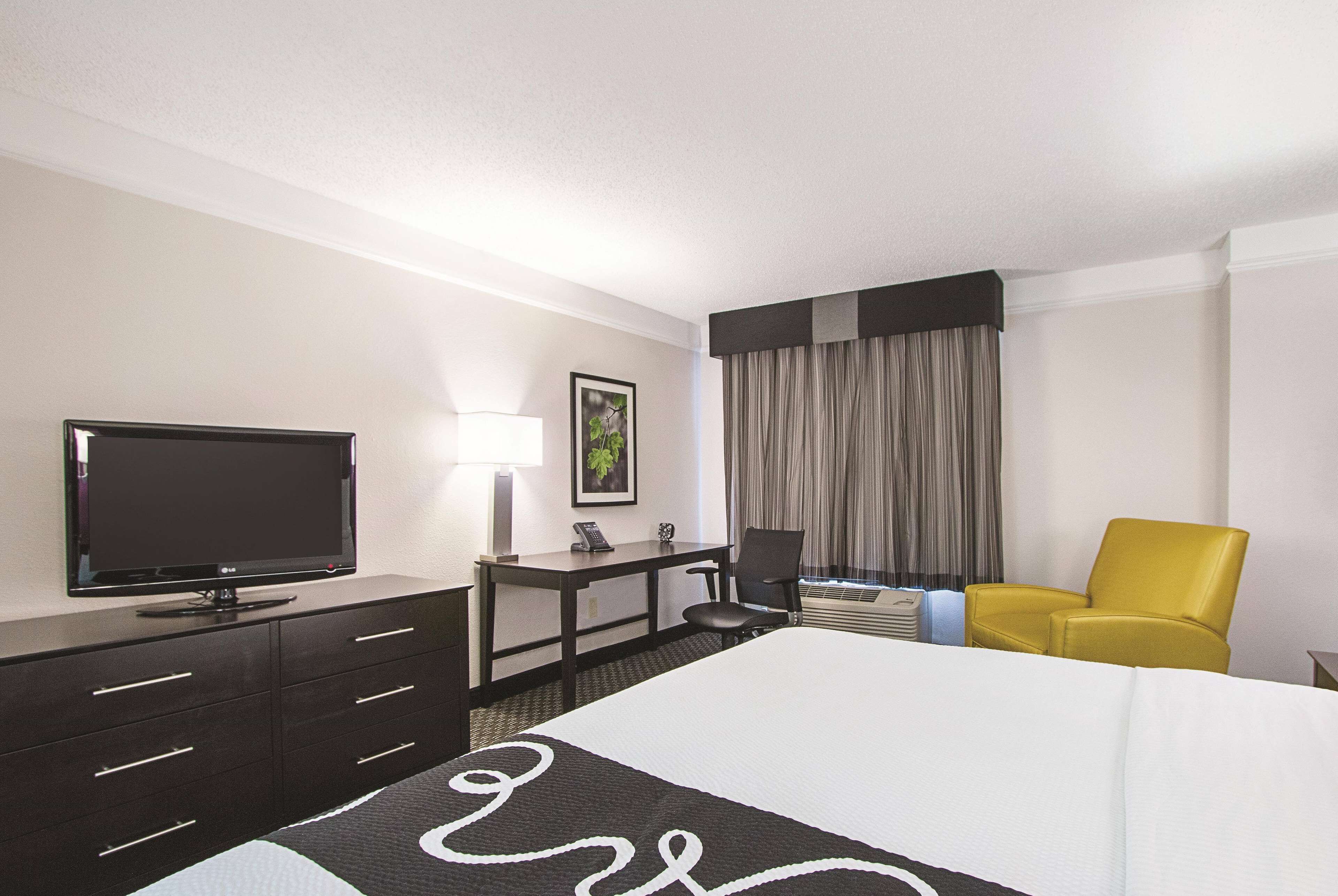 A hotel room with a bed, a desk with a computer, a television, a chair, and a window with curtains.