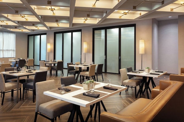 A modern hotel dining area with multiple tables and chairs, large windows, and contemporary lighting fixtures.
