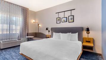 Camera d'albergo con un letto grande, due comodini con lampade, un divanetto, una mensola a parete e una finestra con tende leggere.