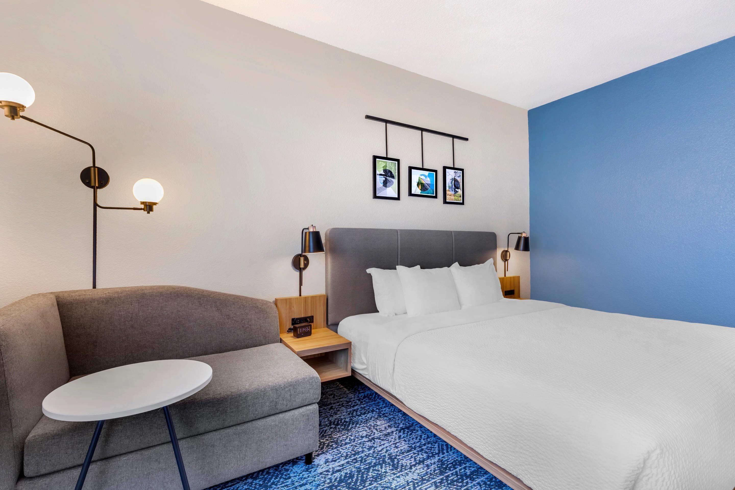 A hotel room with a bed, a sofa, a small table, a nightstand, a lamp, and three framed pictures on the wall.