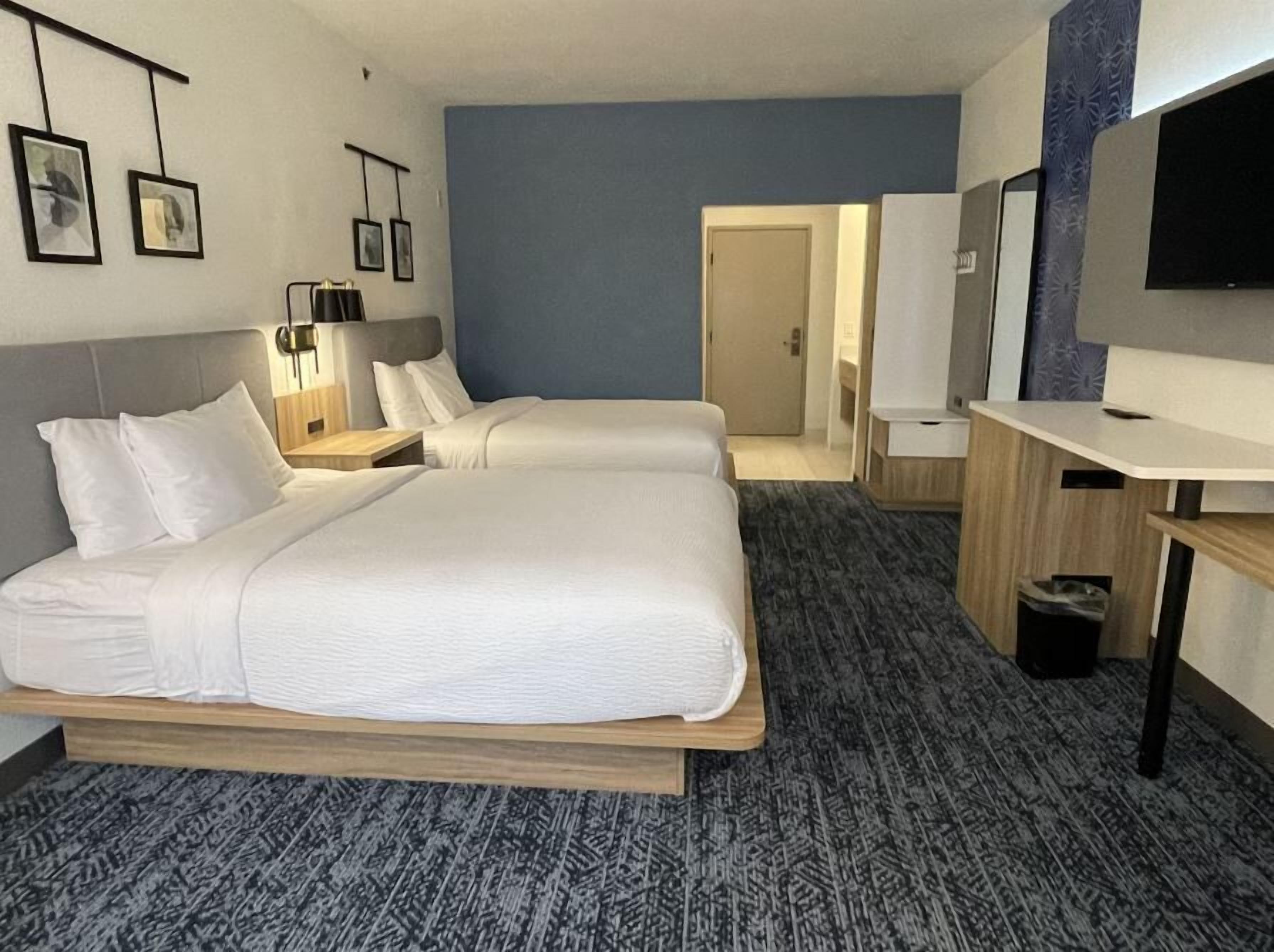 A hotel room with two beds, a desk, a TV, and a door leading to a bathroom.
