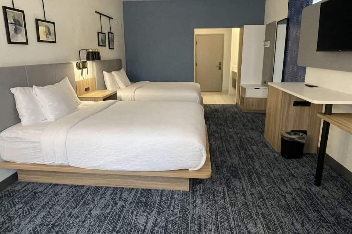 Habitación de hotel con dos camas, un escritorio, un televisor y una puerta que da al baño.
