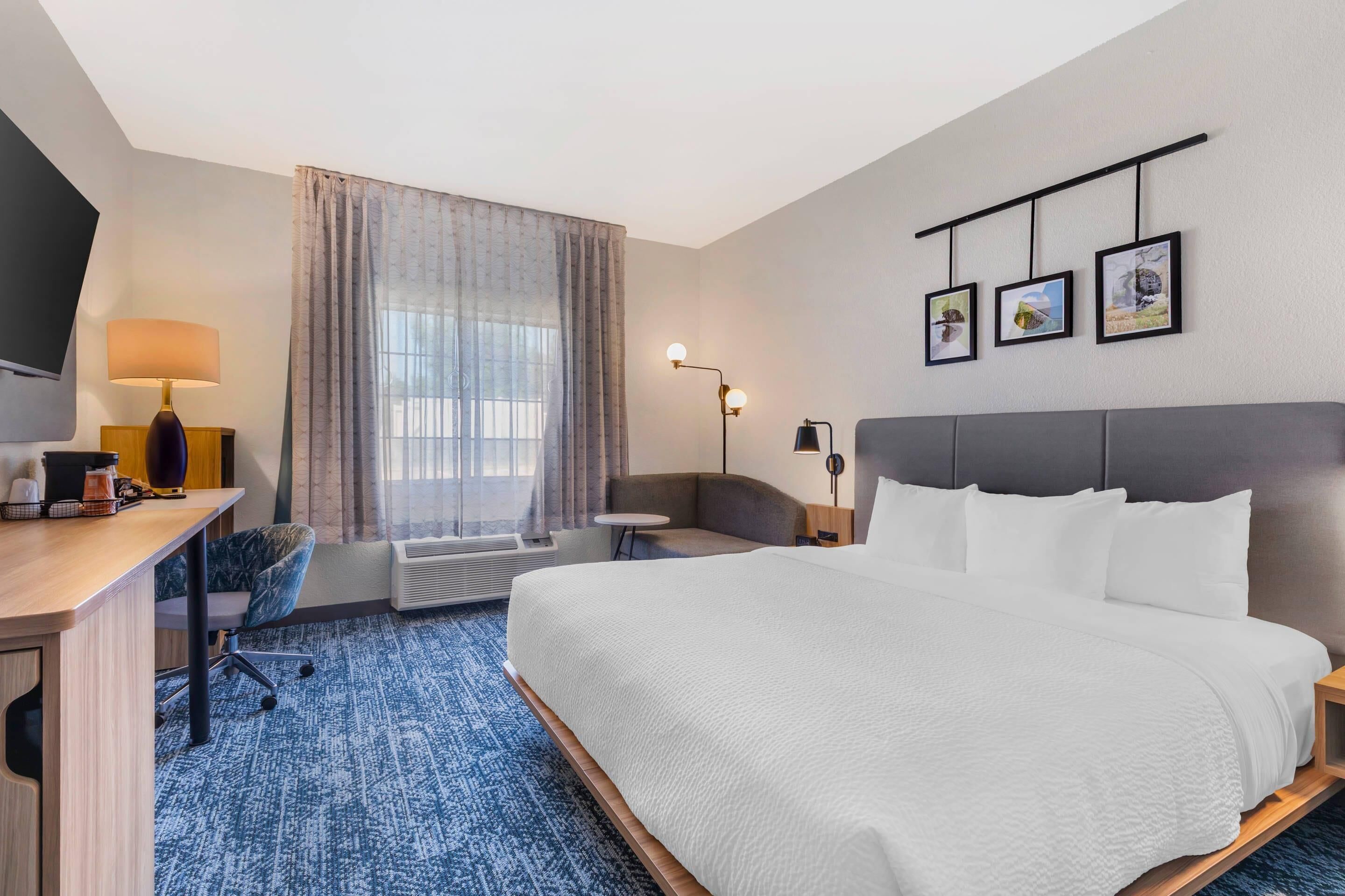 A hotel room with a large bed, a desk with a lamp, a chair, a small table, and a TV. There are three framed pictures on the wall.