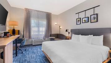 A hotel room with a large bed, a desk with a lamp, a chair, a small table, and a TV. There are three framed pictures on the wall.