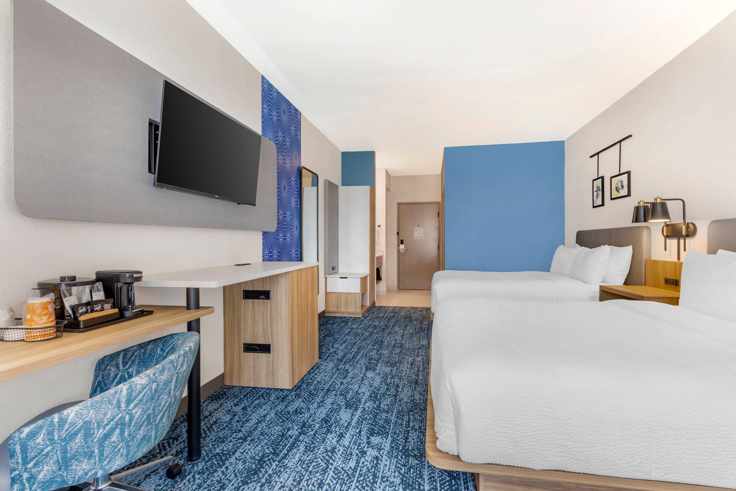 A hotel room with a bed, a desk with a flat-screen TV, a seating area with a chair, and a bathroom visible through an open door.