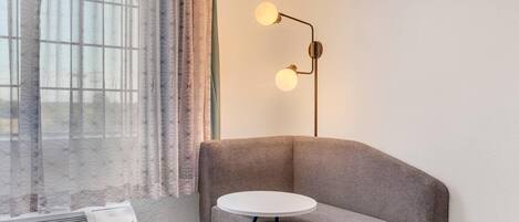 A corner room with a grey sofa, a small round table, a wall-mounted lamp, and a window with sheer curtains.