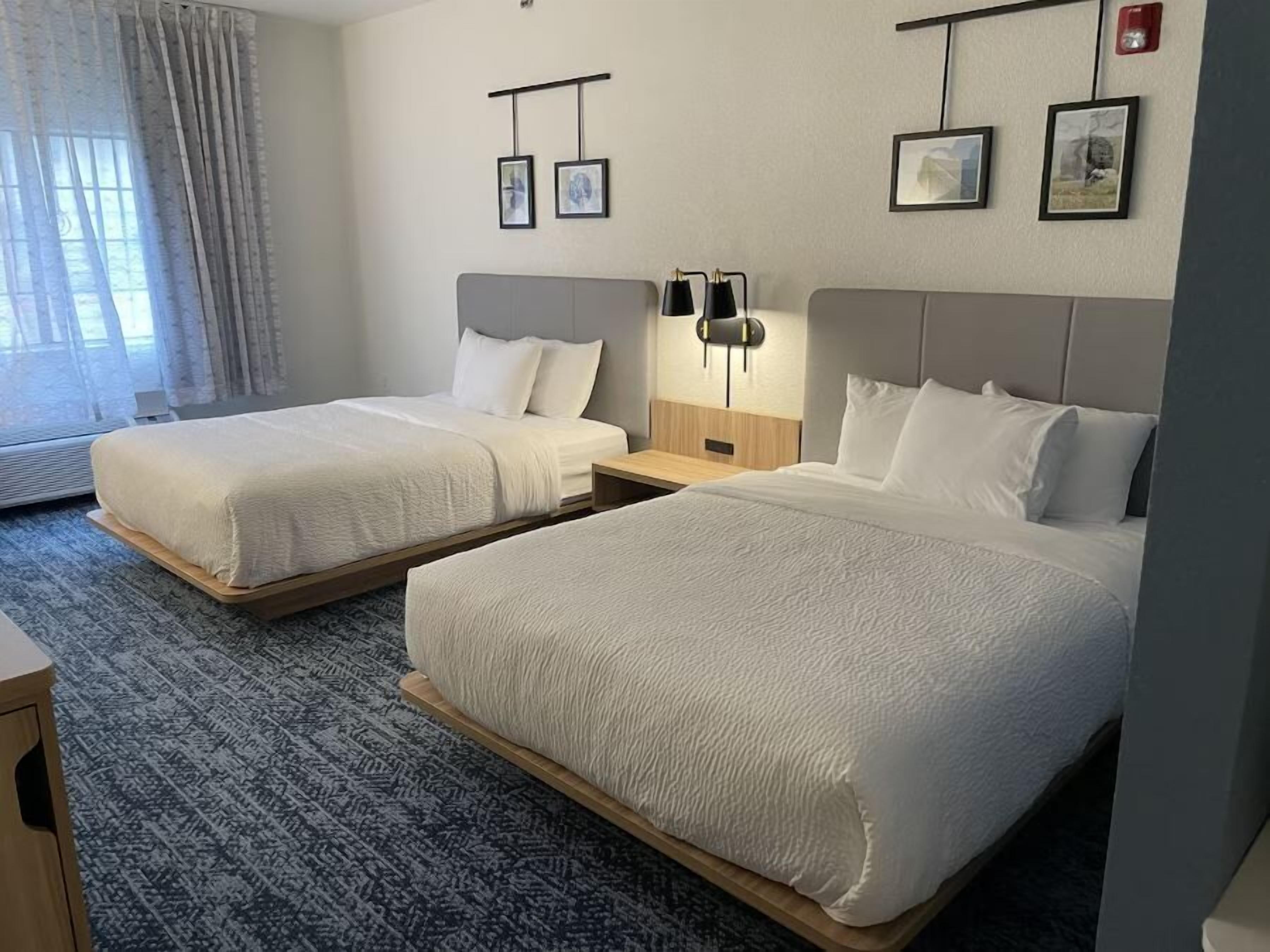 A hotel room with two beds, a bedside table, a lamp, and framed pictures on the wall.