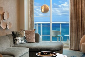 Un salon moderne avec un canapé gris, des coussins décoratifs, une table basse ronde sur laquelle se trouve un bol d’œufs, et une vue sur l’océan par de grandes fenêtres.