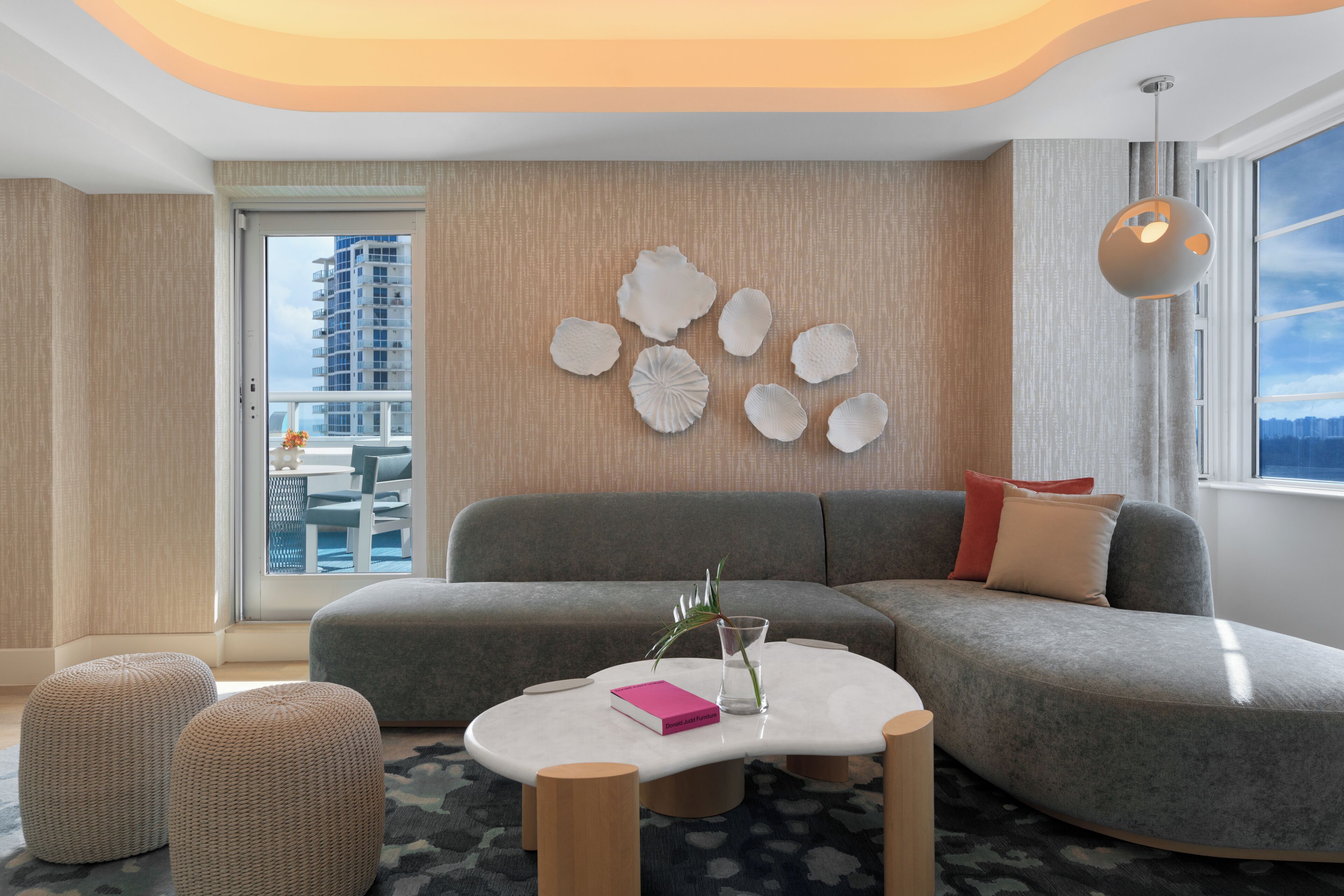 Un salon moderne avec un canapé gris, des poufs, une table basse blanche et une vue sur la ville à travers de grandes fenêtres.