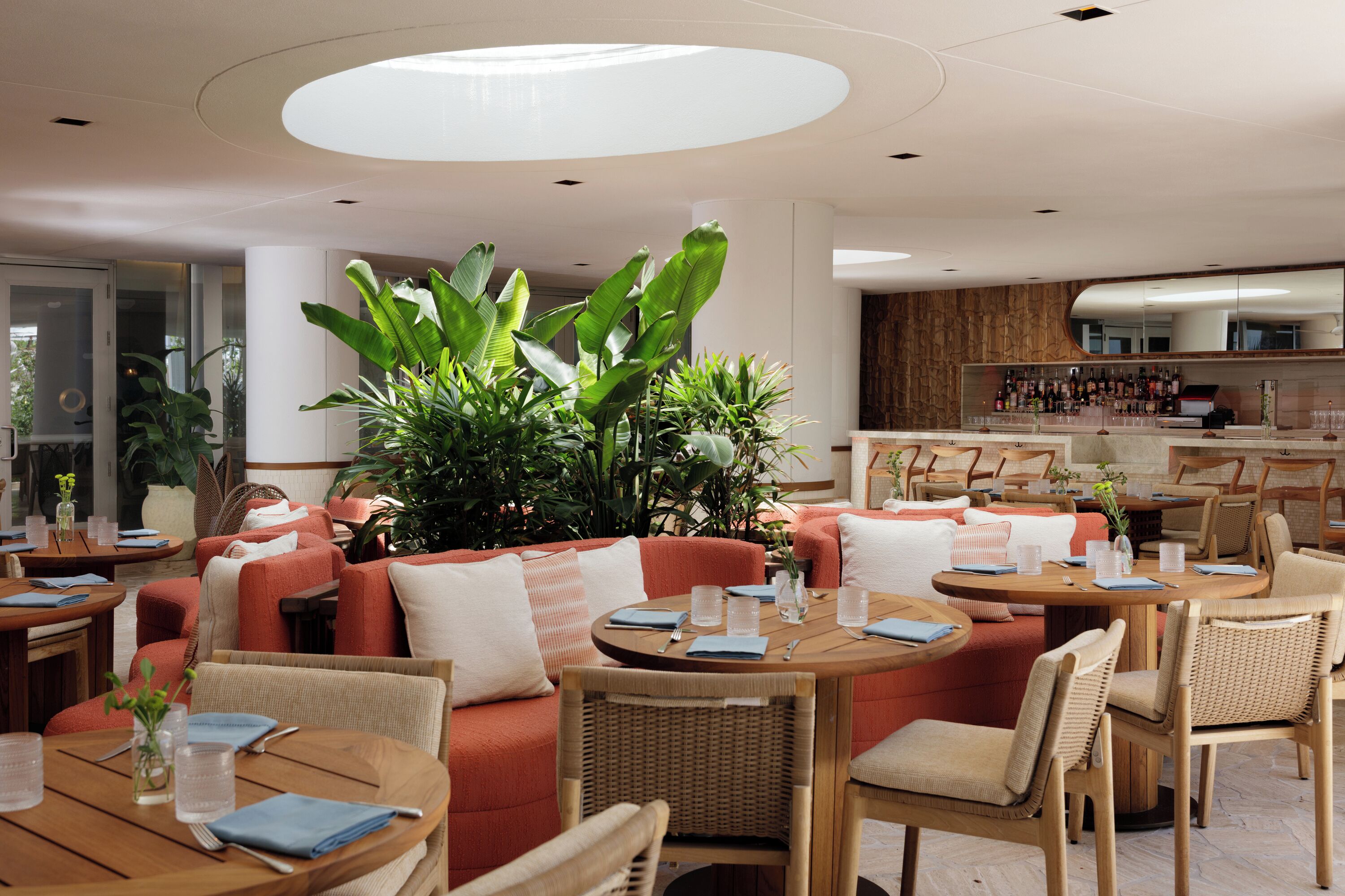 A modern restaurant with round tables and wicker chairs, a bar area, and large green plants.