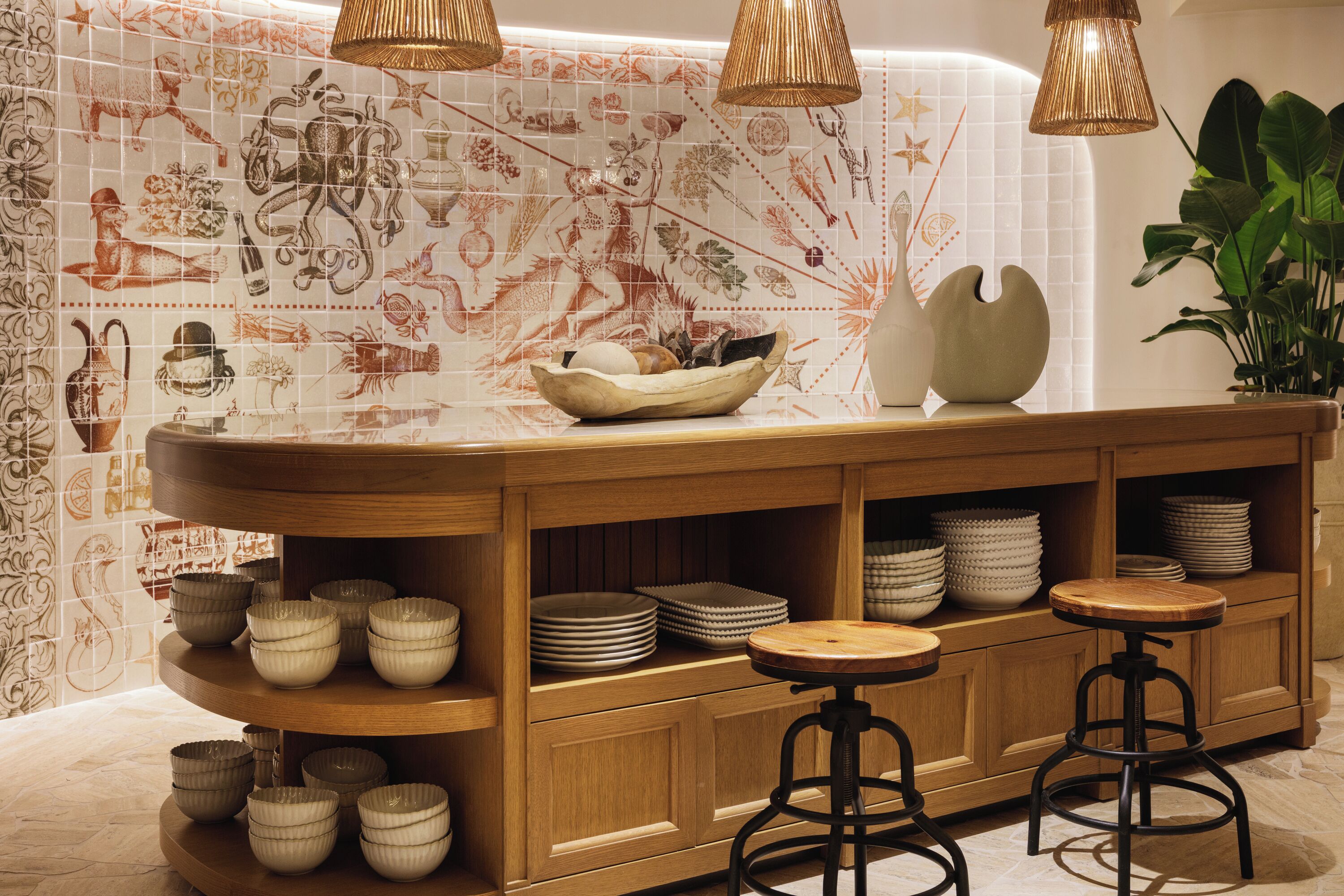 Une cuisine avec un mur recouvert de carreaux décoratifs, des étagères en bois où sont posés des bols et des assiettes, et deux tabourets de bar.