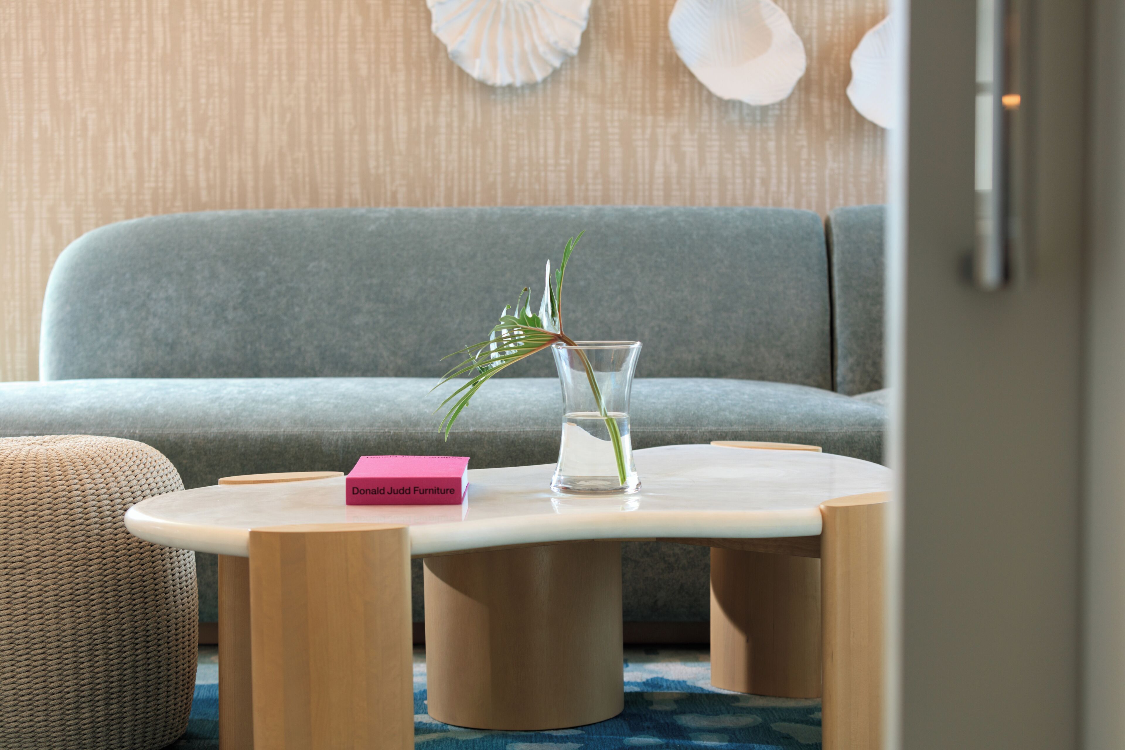 Ein modernes Wohnzimmer mit einem grauen Sofa, einem runden Couchtisch mit einer Glasvase und einer Pflanze, sowie einer pinkfarbenen Box mit der Aufschrift 'Donald Judd Furniture'.