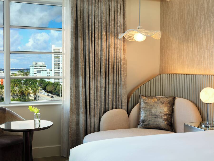 A hotel room with a bed, a round table with a vase, a chair, a view of the cityscape through a window, and a hanging light fixture.