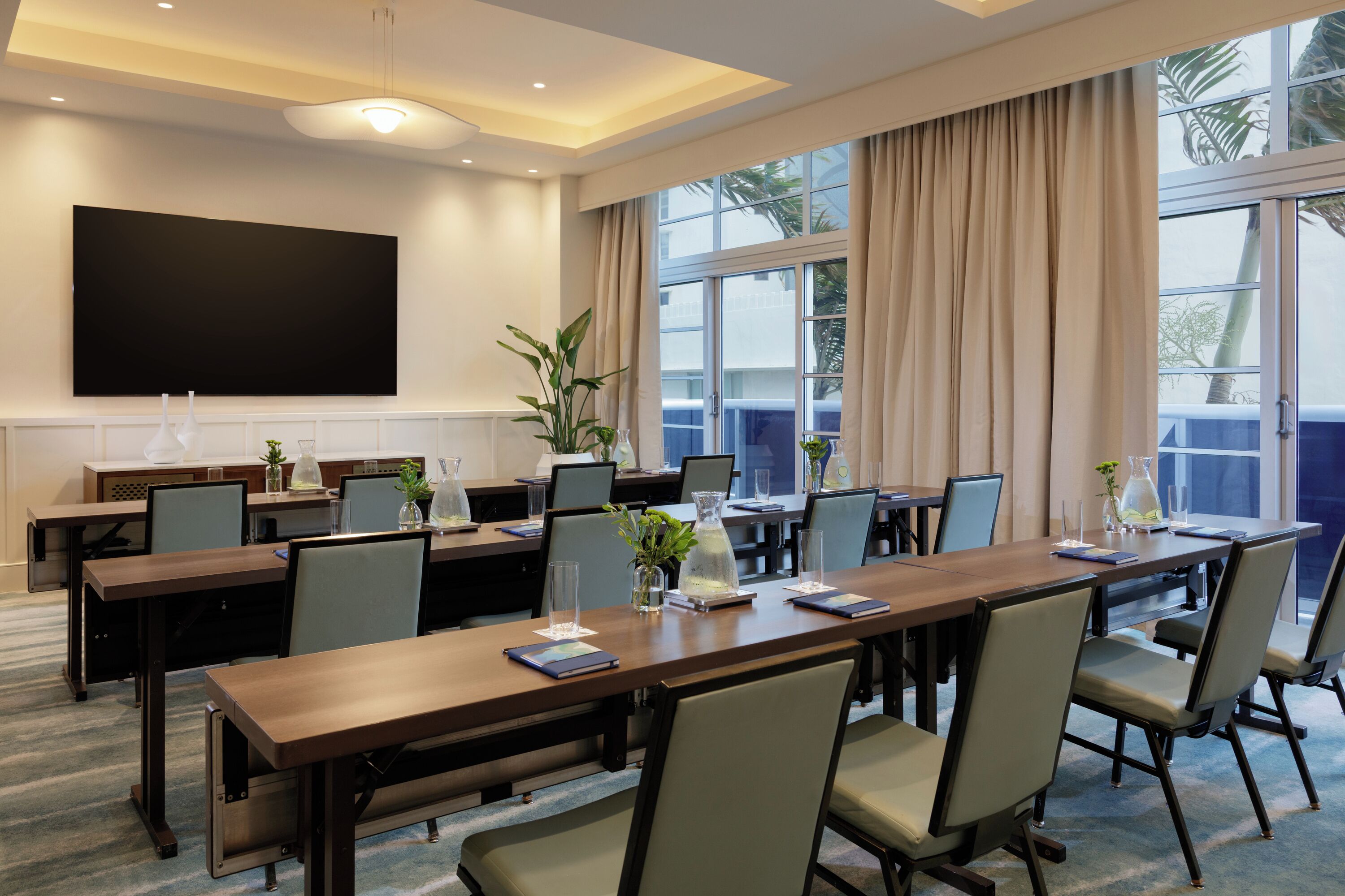 A conference room with a long table, chairs, a flat-screen TV, and large windows with curtains.