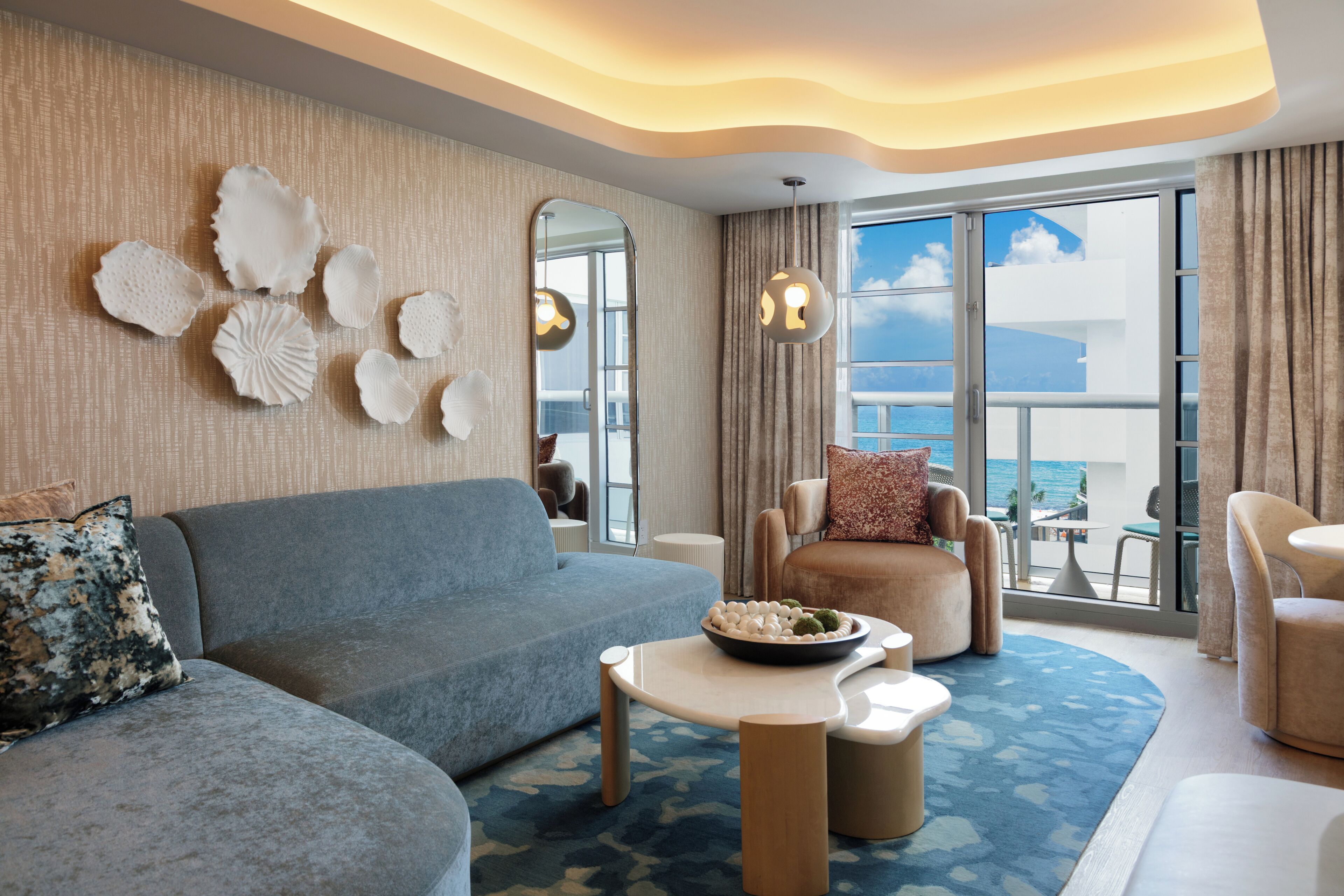 A modern hotel room with a large window, a blue sofa, a round coffee table, and a decorative wall with shells.