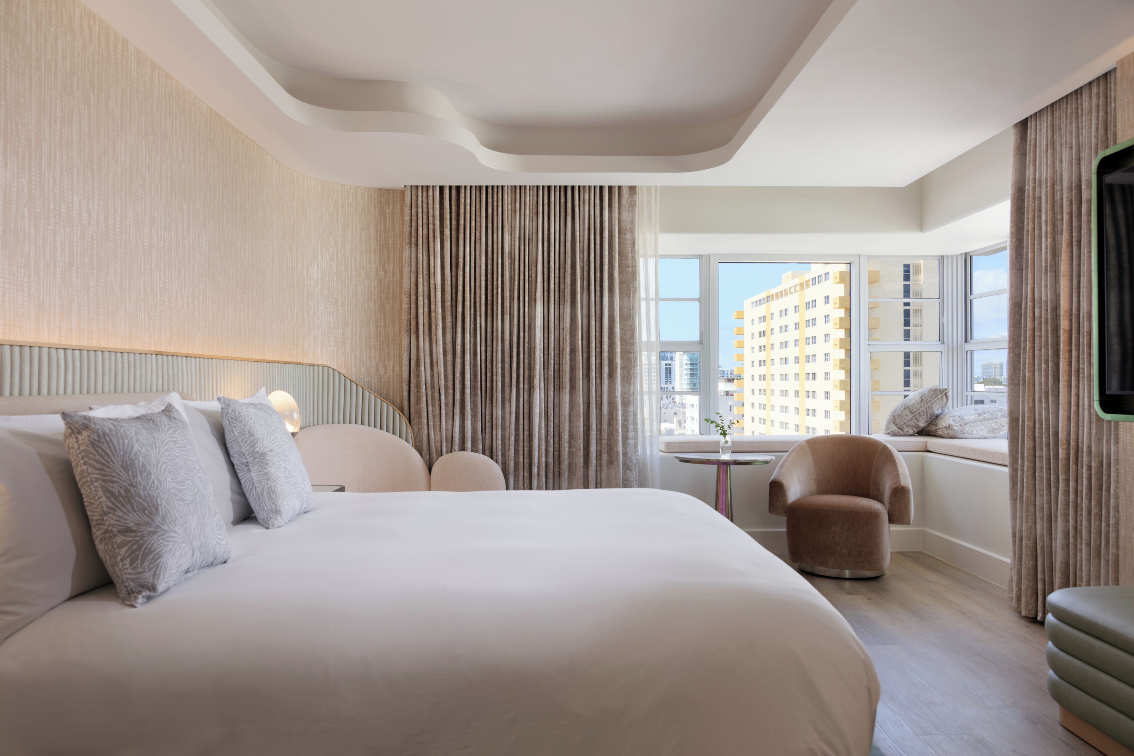 Een moderne hotelkamer met een groot bed, een zithoek met een stoel en een tafeltje, een raam met uitzicht op gebouwen en een aan de muur bevestigde televisie.