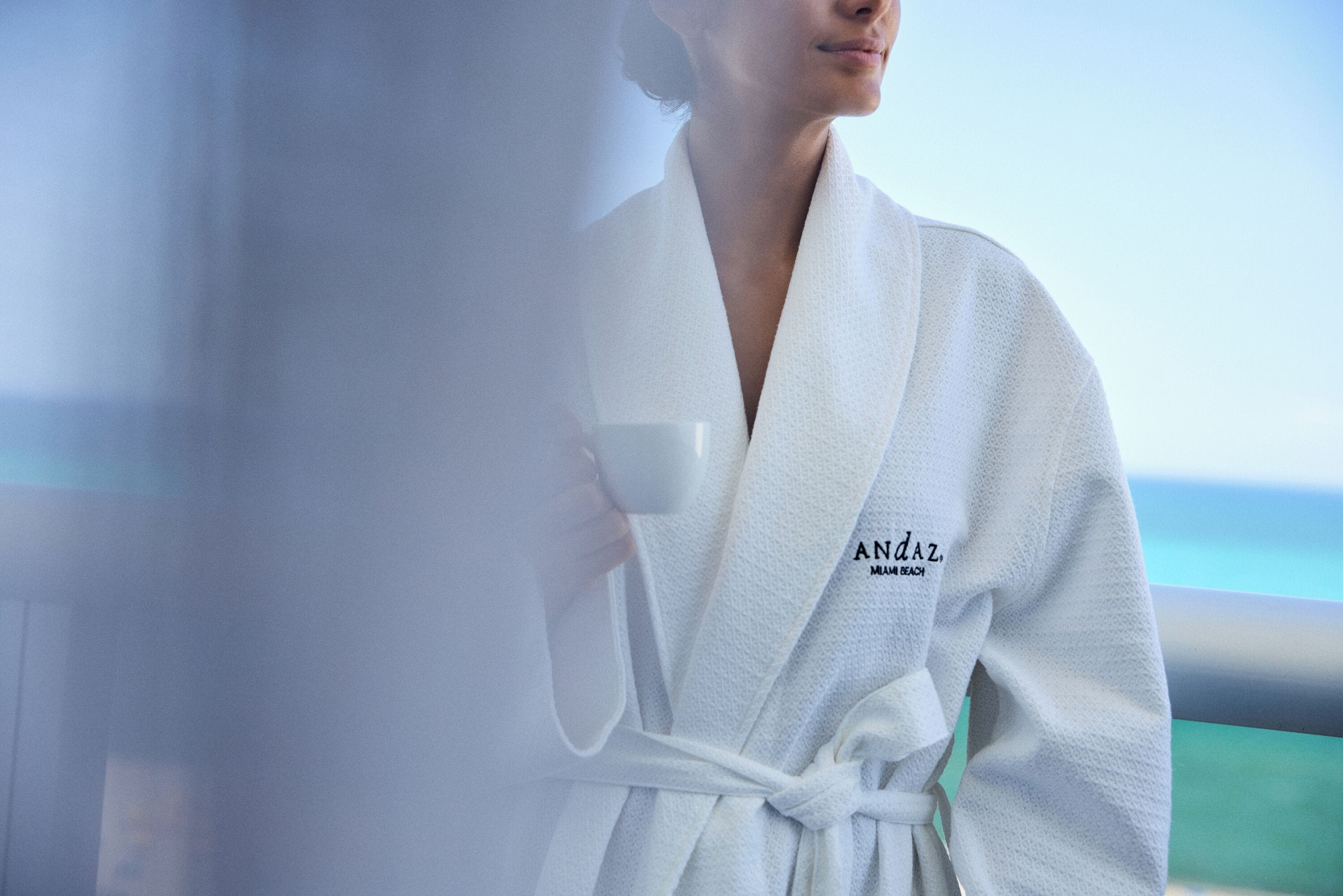 Eine Person in einem weißen Bademantel mit der Aufschrift 'ANZ' und 'MIAMI BEACH', stehend am Fenster mit Blick aufs Meer.