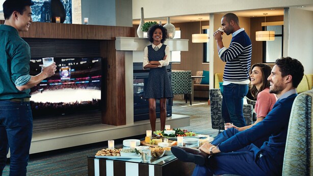 Uma sala de estar moderna com televisão de tela plana, uma mesa de centro com petiscos e um espaço de estar com sofá e poltronas.