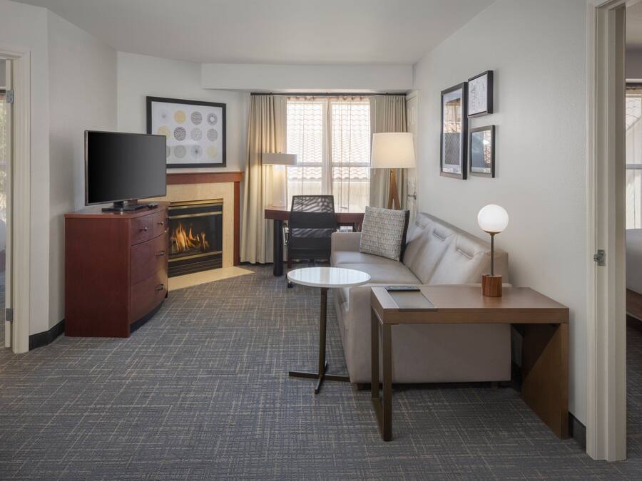 A hotel room with a flat-screen TV, a sofa, a small table, a fireplace, and a window with curtains.