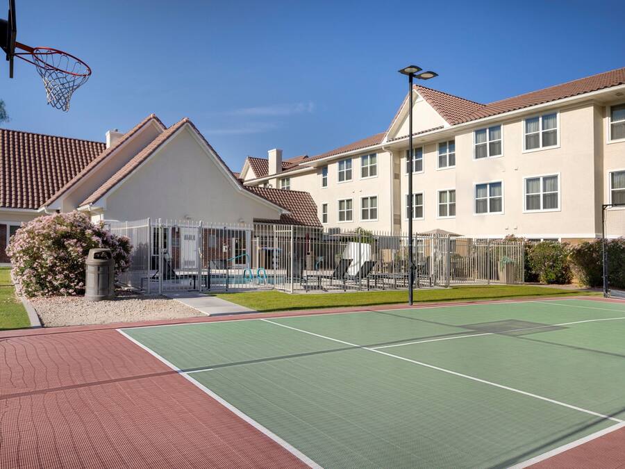 Uma quadra de basquete ao ar livre com superfície verde, cercada por uma grade, e adjacente a um edifício com telhado de telhas vermelhas.
