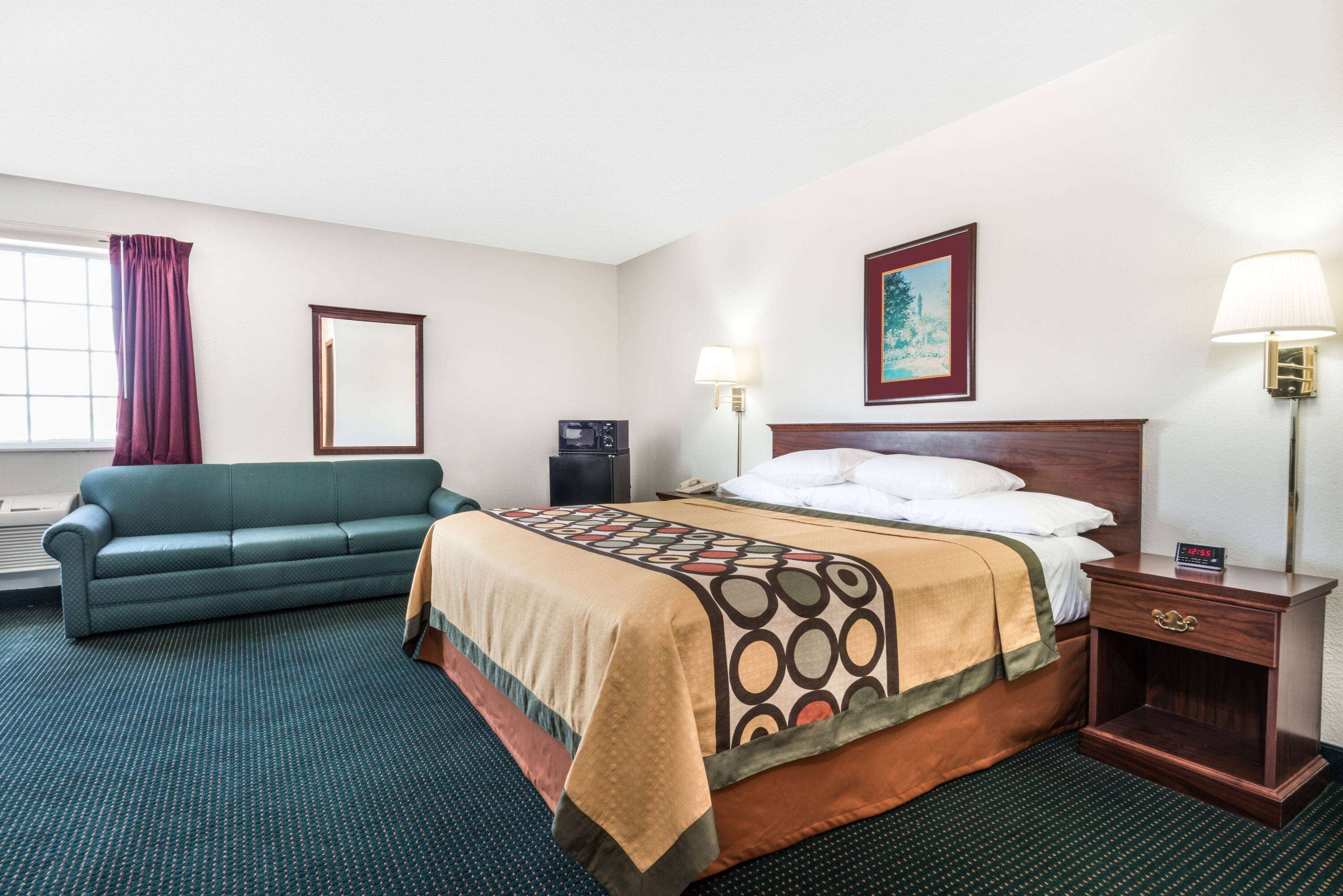 Una habitación de hotel con una cama, un sofá verde, una mesita de noche con una lámpara y un cuadro en la pared.