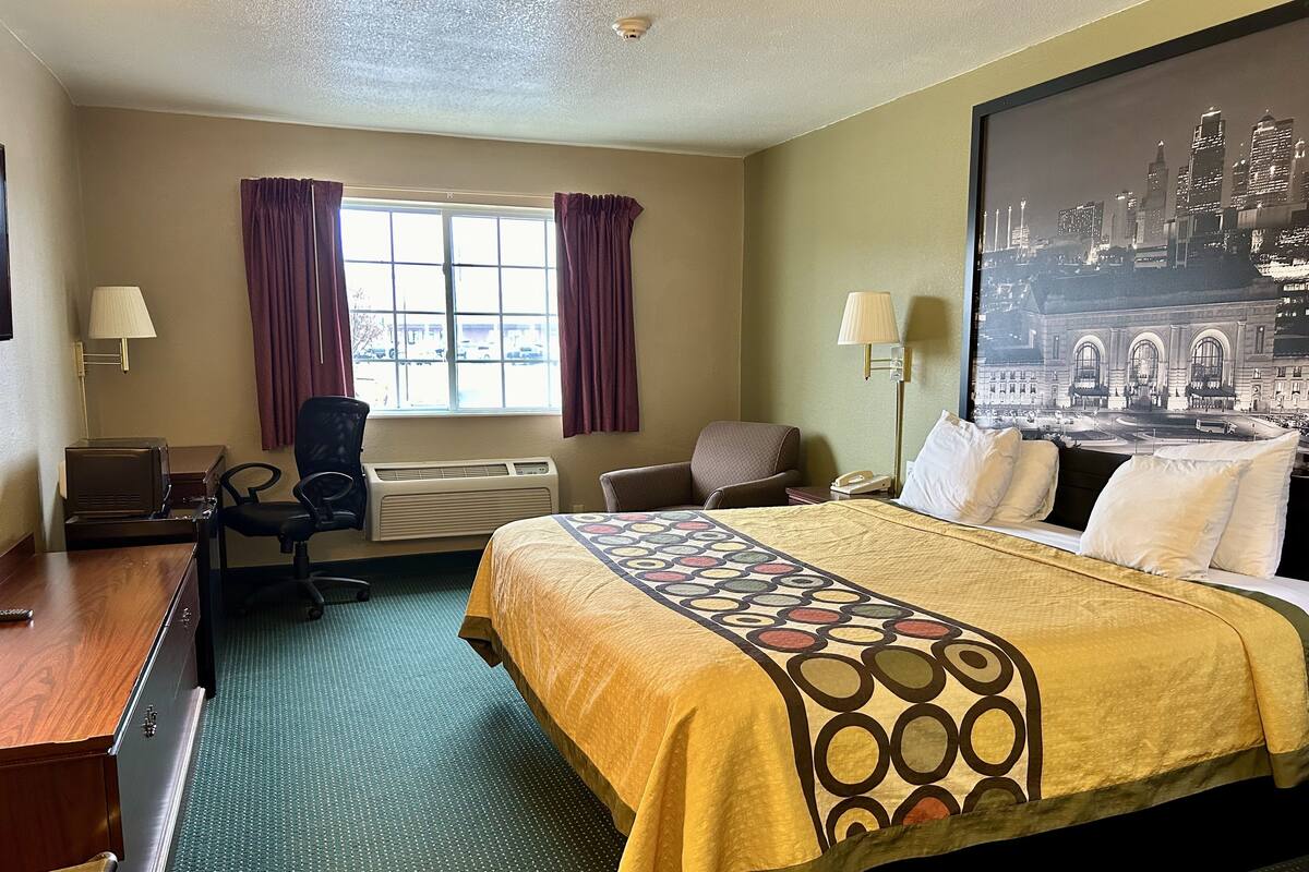 Et hotelværelse med en seng, to stole, et skrivebord, en lampe, en varmeovn og et stort vindue med gardiner.