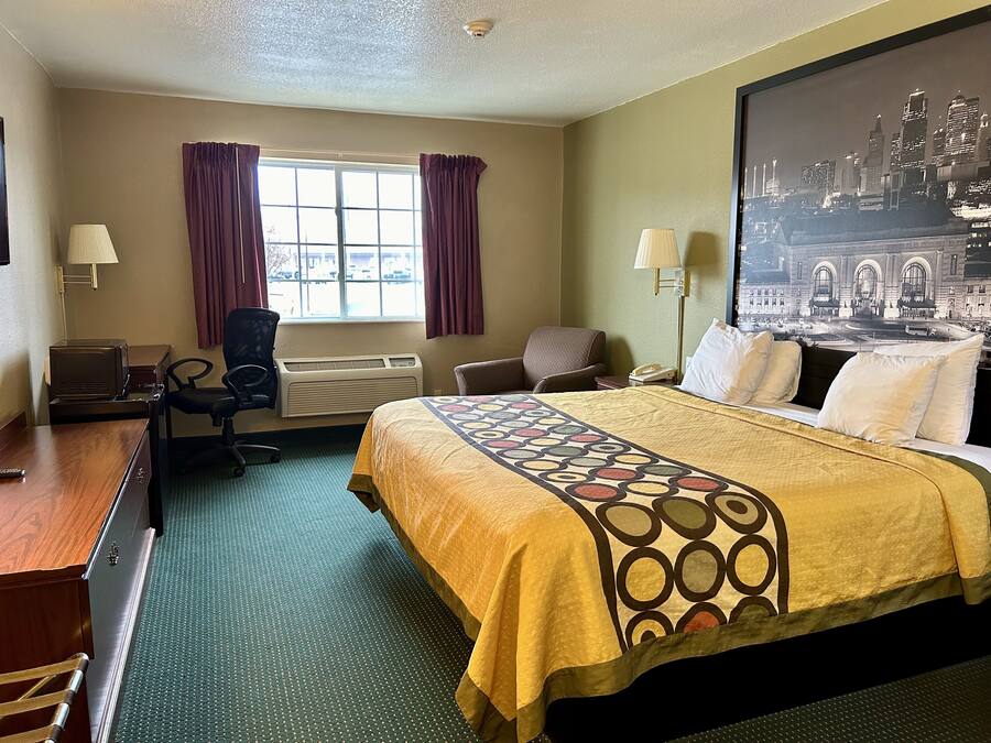 A hotel room with a bed, two chairs, a desk, a lamp, a heater, and a large window with curtains.