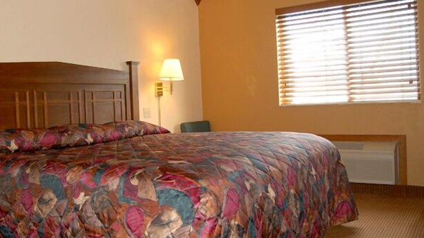 A bedroom with a wooden headboard, a patterned bedspread, a bedside lamp, a chair, and a window with blinds.