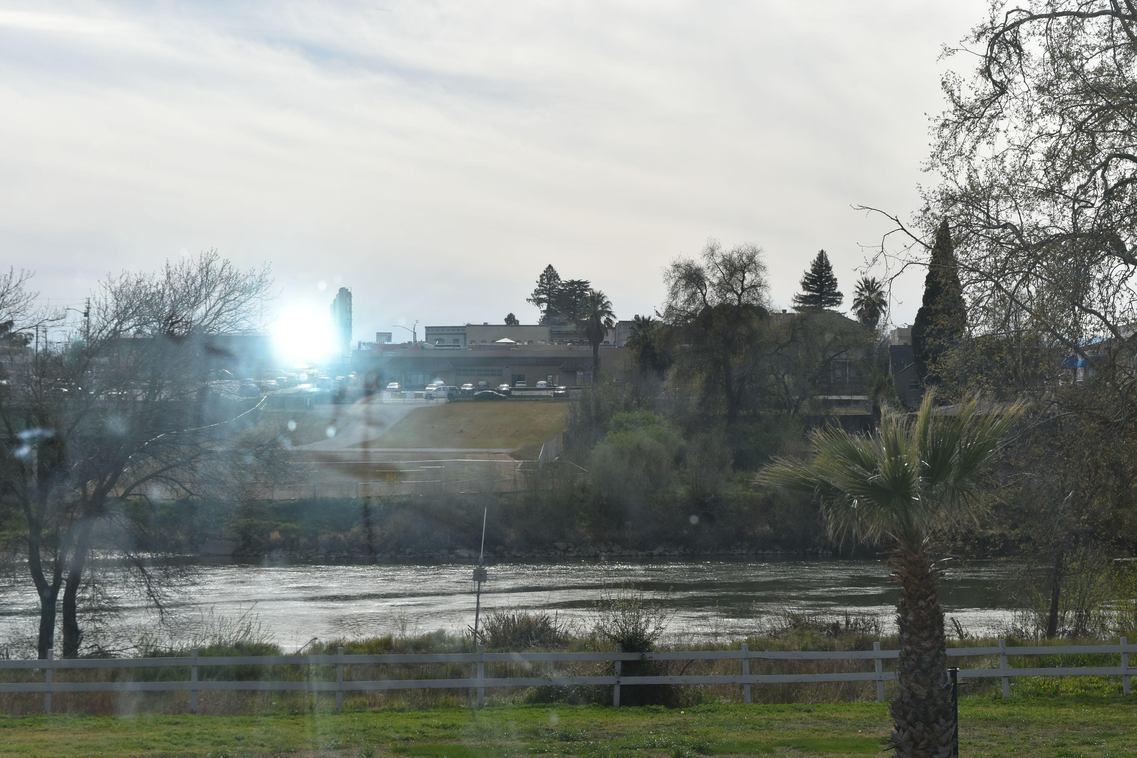 A river with a white fence in the foreground, trees, and buildings in the background.