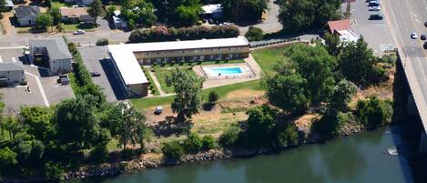 An aerial view of a building with a swimming pool, surrounded by greenery and a river.