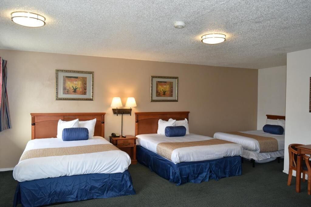 A hotel room with three single beds, each with a wooden headboard, white bedding, and blue accents. There are two wall-mounted lamps and two ceiling lights.