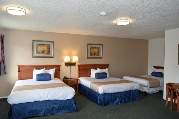 A hotel room with three single beds, each with a wooden headboard, white bedding, and blue accents. There are two wall-mounted lamps and two ceiling lights. - Travelodge by Wyndham Red Bluff South of Redding (Red Bluff)