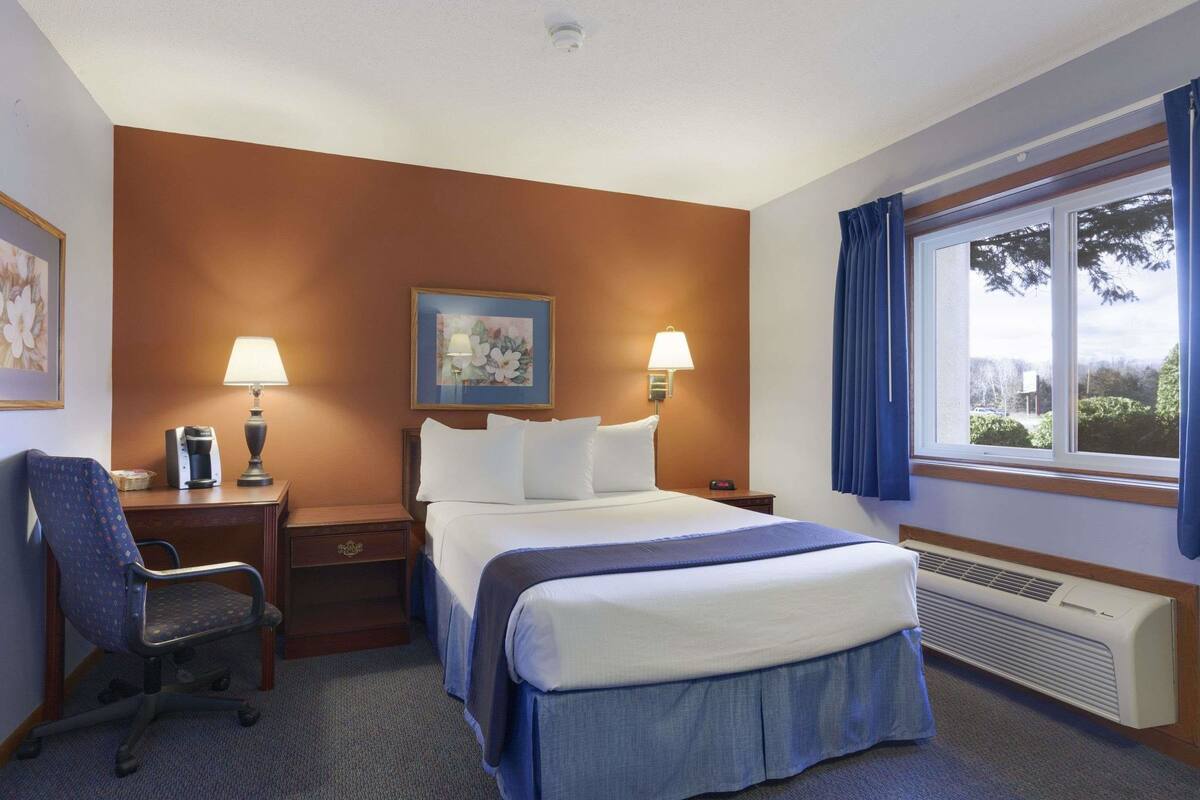A hotel room with a bed, two bedside lamps, a desk with a chair, a framed picture on the wall, and a window with curtains.