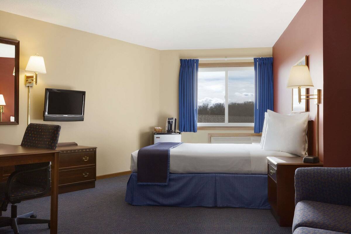 A hotel room with a bed, a desk with a chair, a television, a nightstand, and a window with curtains.