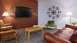 Ein Zimmer mit einem braunen Ledersofa, gemusterten Sessel und einem Holztisch. Zwei Lampen sorgen für Licht. An der Wand ist ein Flachbildfernseher angebracht, darüber befindet sich eine dekorative Metallskulptur.