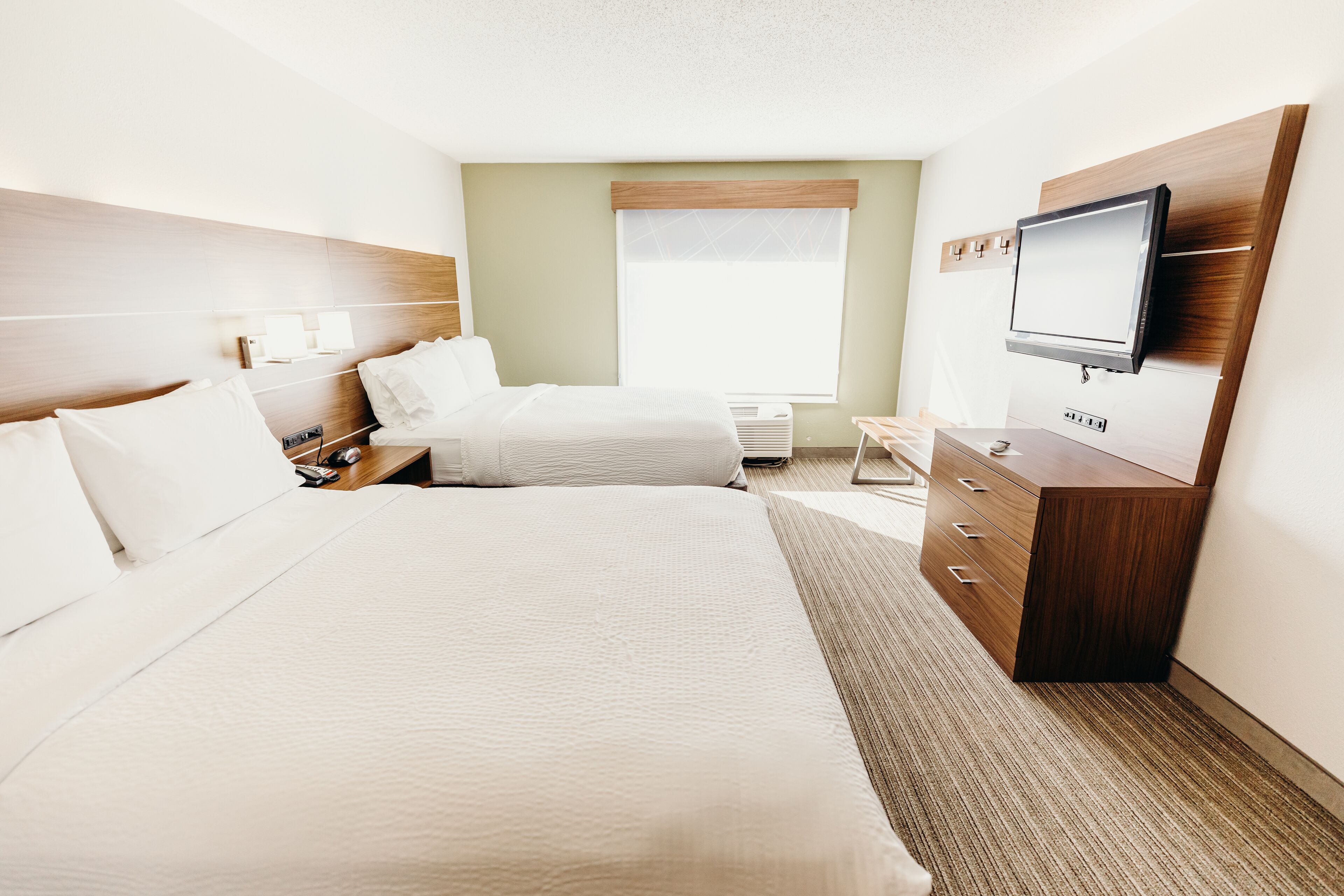 A hotel room with a large bed, a smaller bed, a TV mounted on the wall, a desk, and a window with blinds.