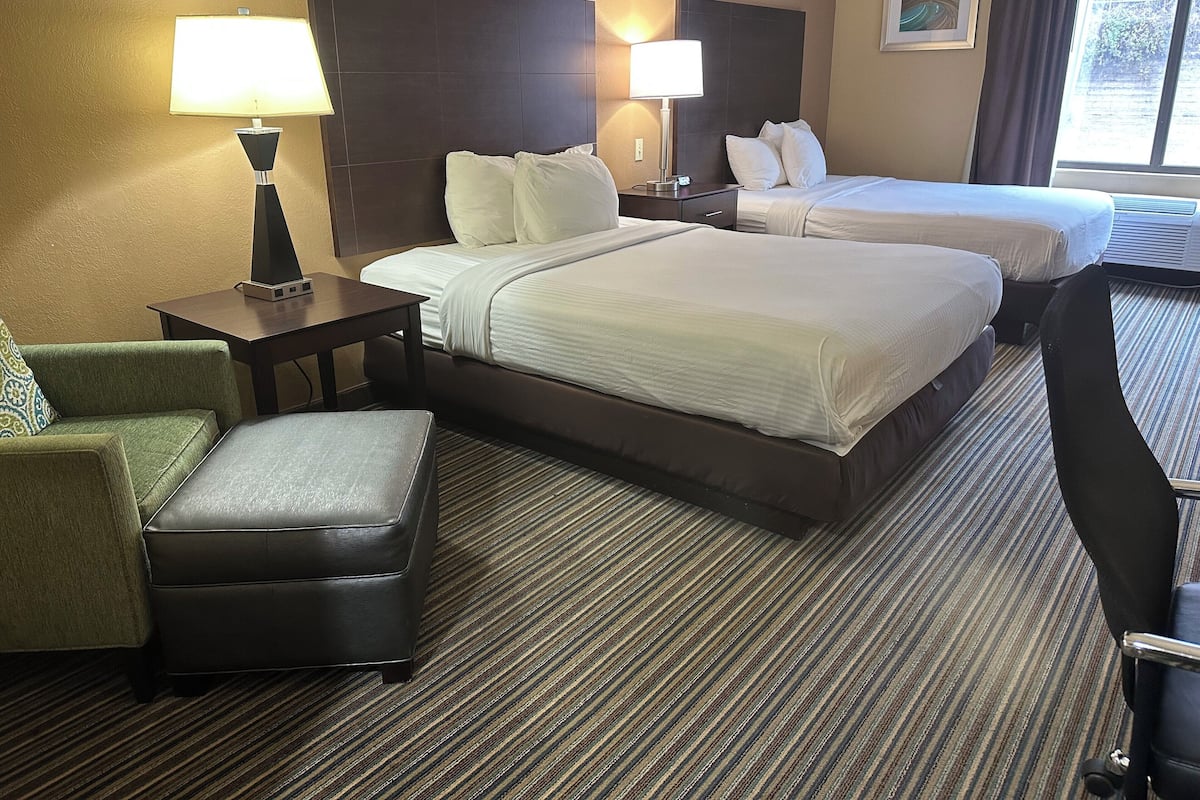 A hotel room with two beds, a chair, a small table, and a lamp.