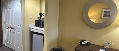 Habitación de hotel con kitchenette, un refrigerador pequeño, un basurero, un escritorio con teléfono y un espejo montado en la pared.